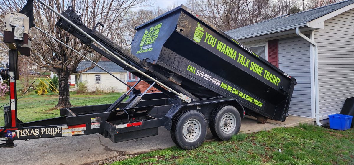 A black Talkin' Trash 12-yard roll-off dumpster on a trailer, being delivered to a home driveway in Marion, NC
